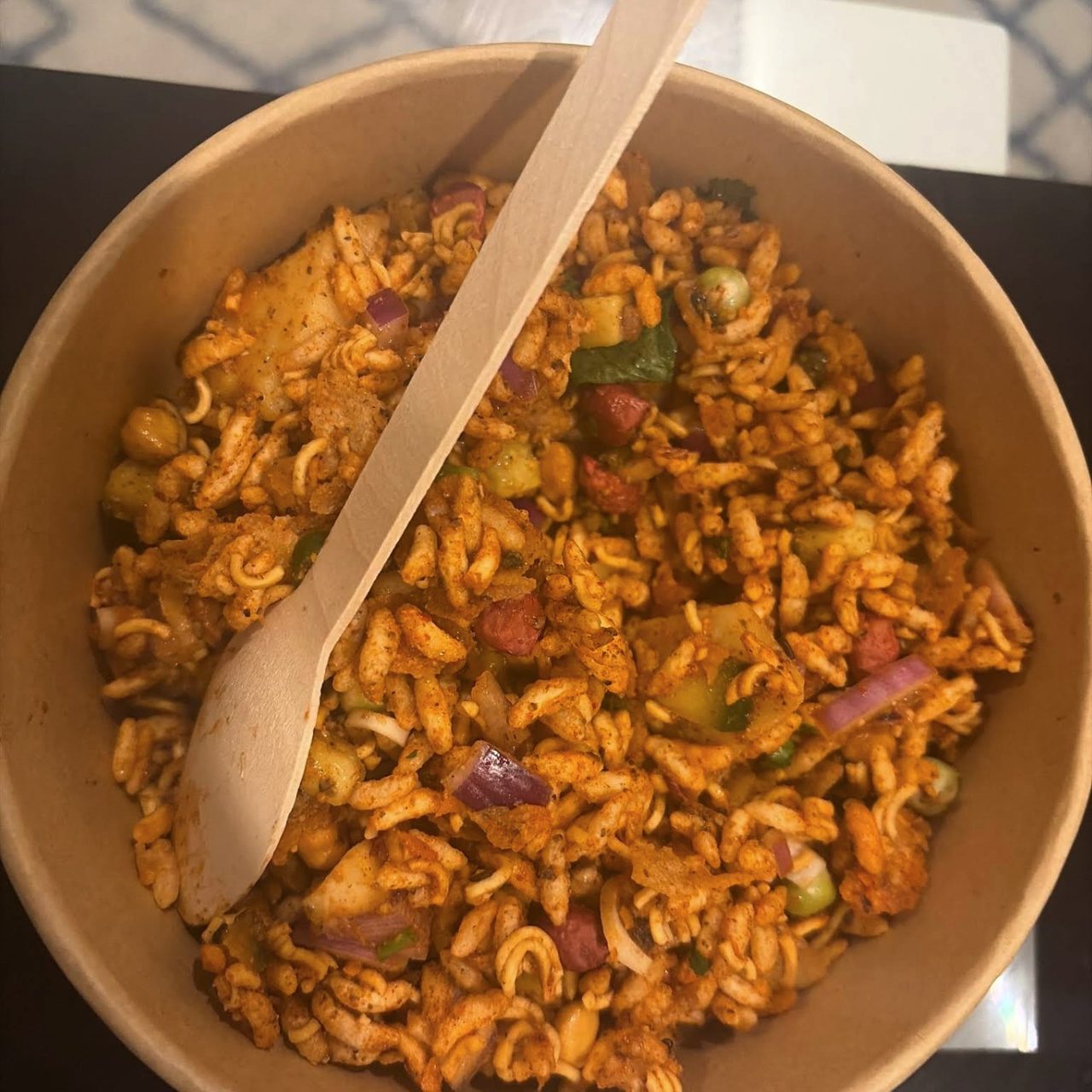 A bowl of Amilo Piro chatpate — puffed rice, noodles, potato and onion in red masala, with a wooden spoon