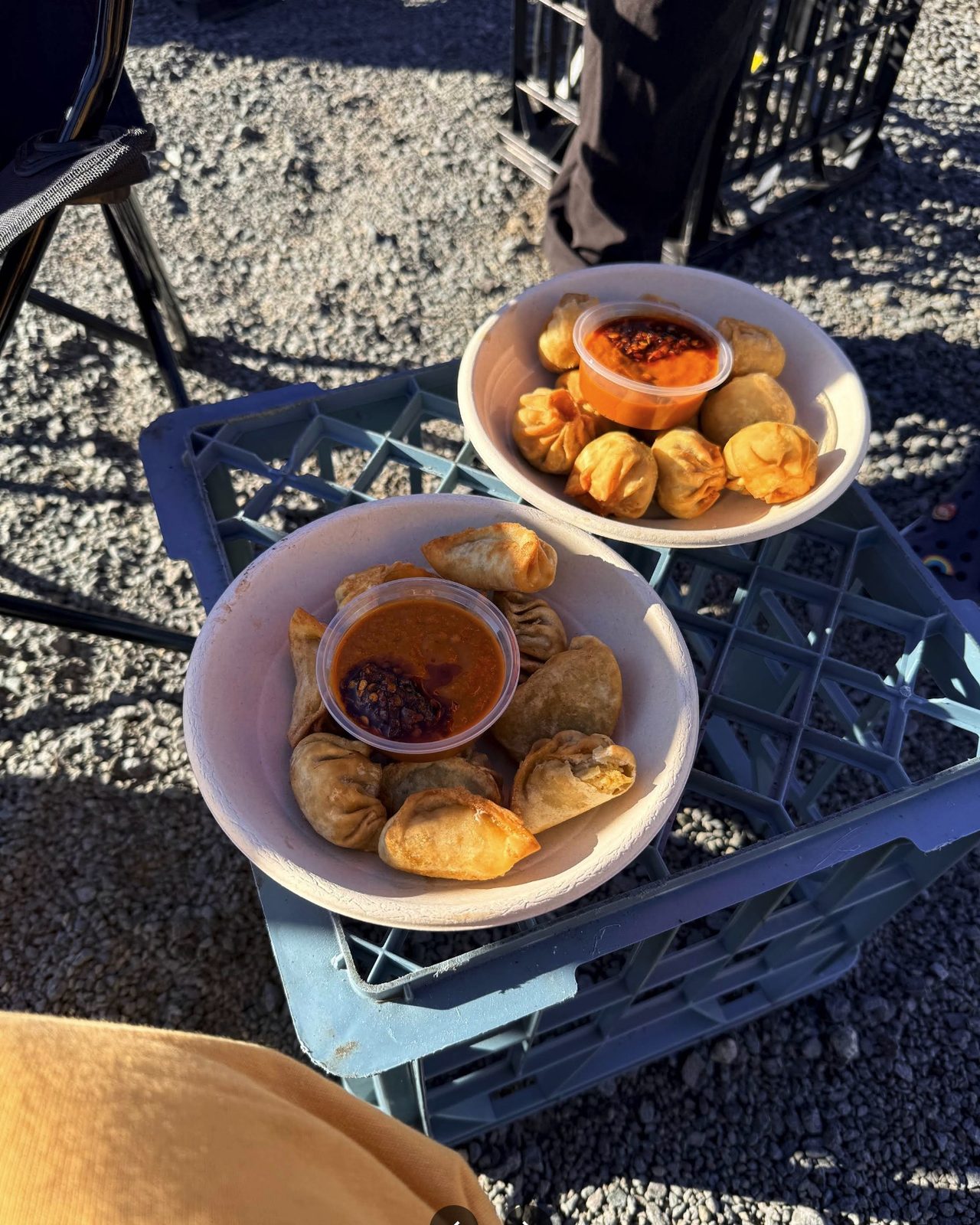 Two bowls of fried Amilo Piro momo with cups of chilli sauce, on a crate in late afternoon sun