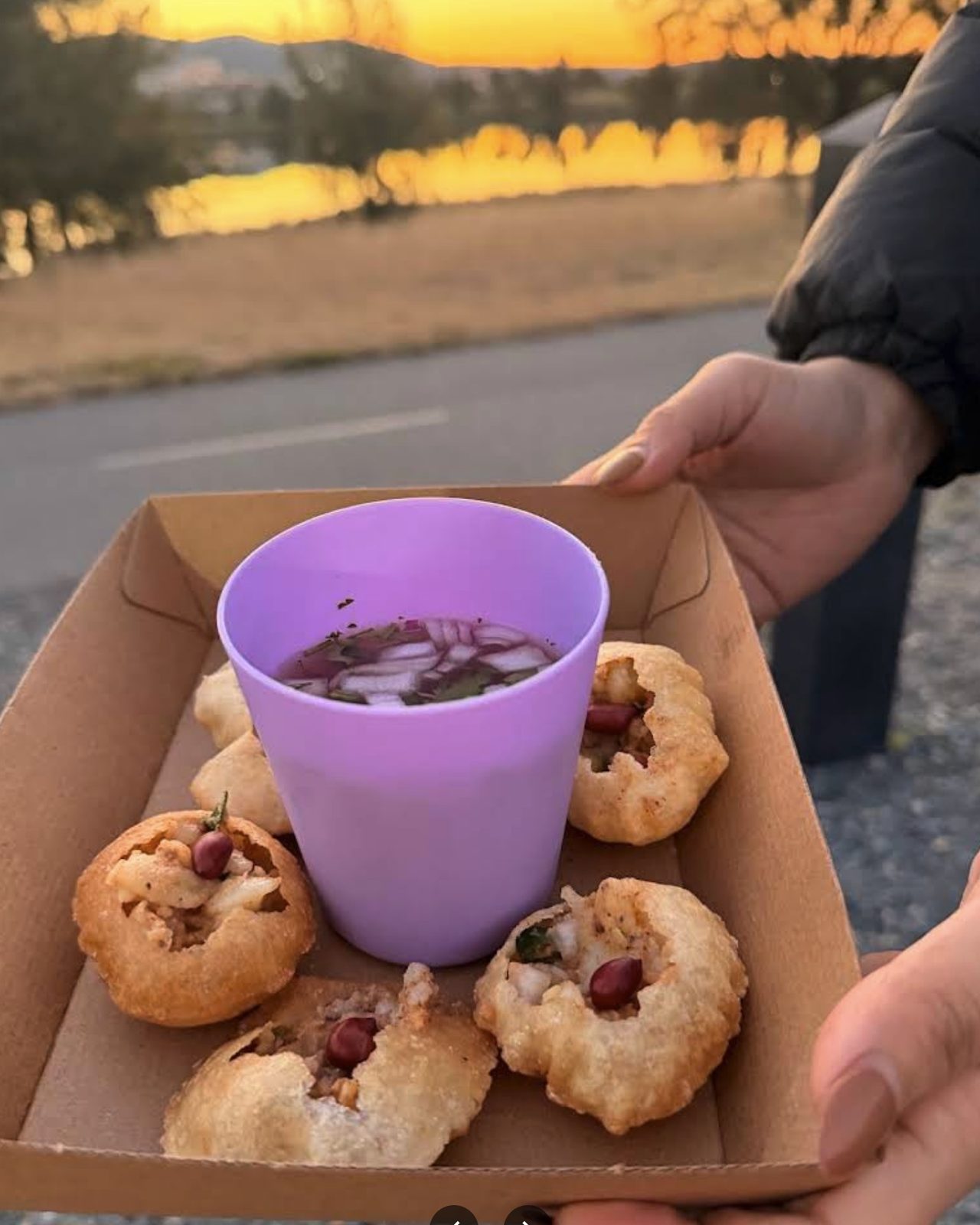 Six Amilo Piro pani puri in a box around a cup of jhol, held up against a sunset