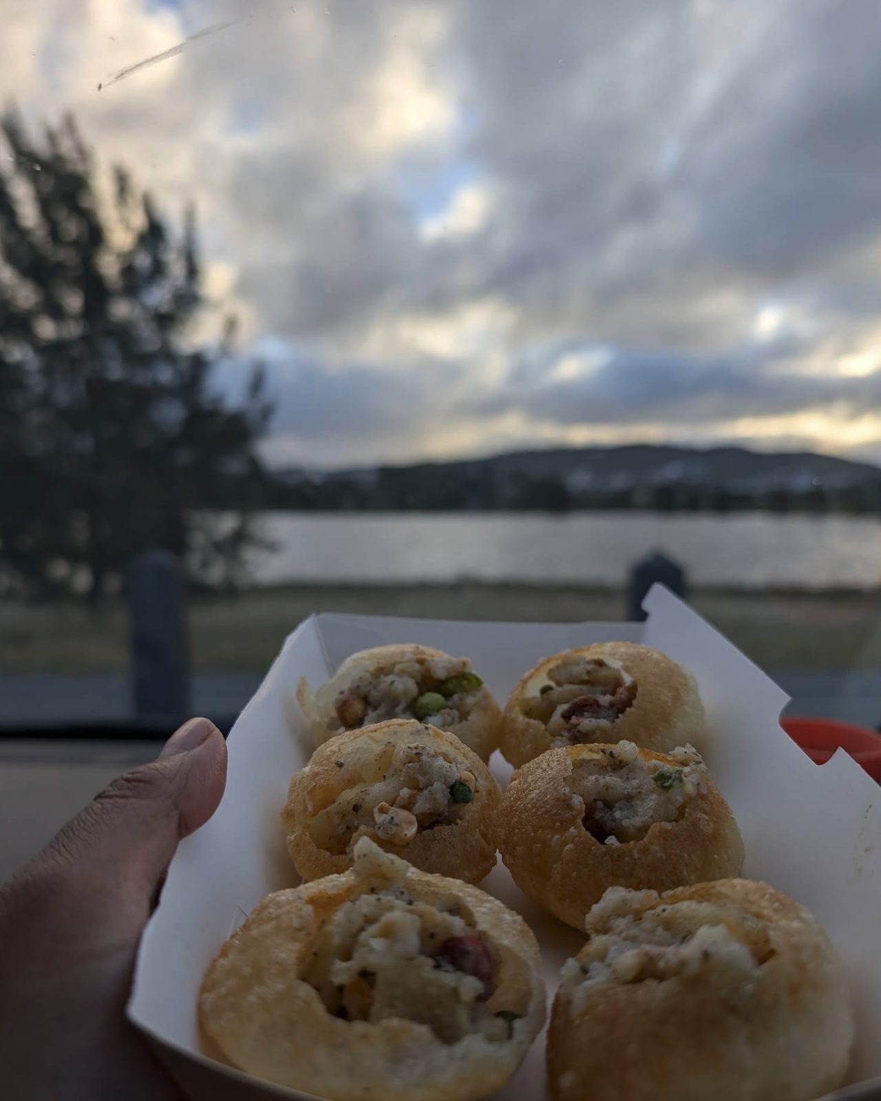 A box of Amilo Piro pani puri and a cup of jhol held up beside the pond at dusk